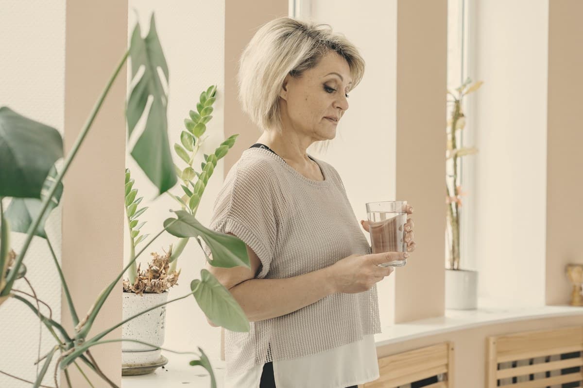 A calm woman holding a glass of water at a bright window, reflecting the daily oral medication routine discussed in the approved GLP-1 pill section.