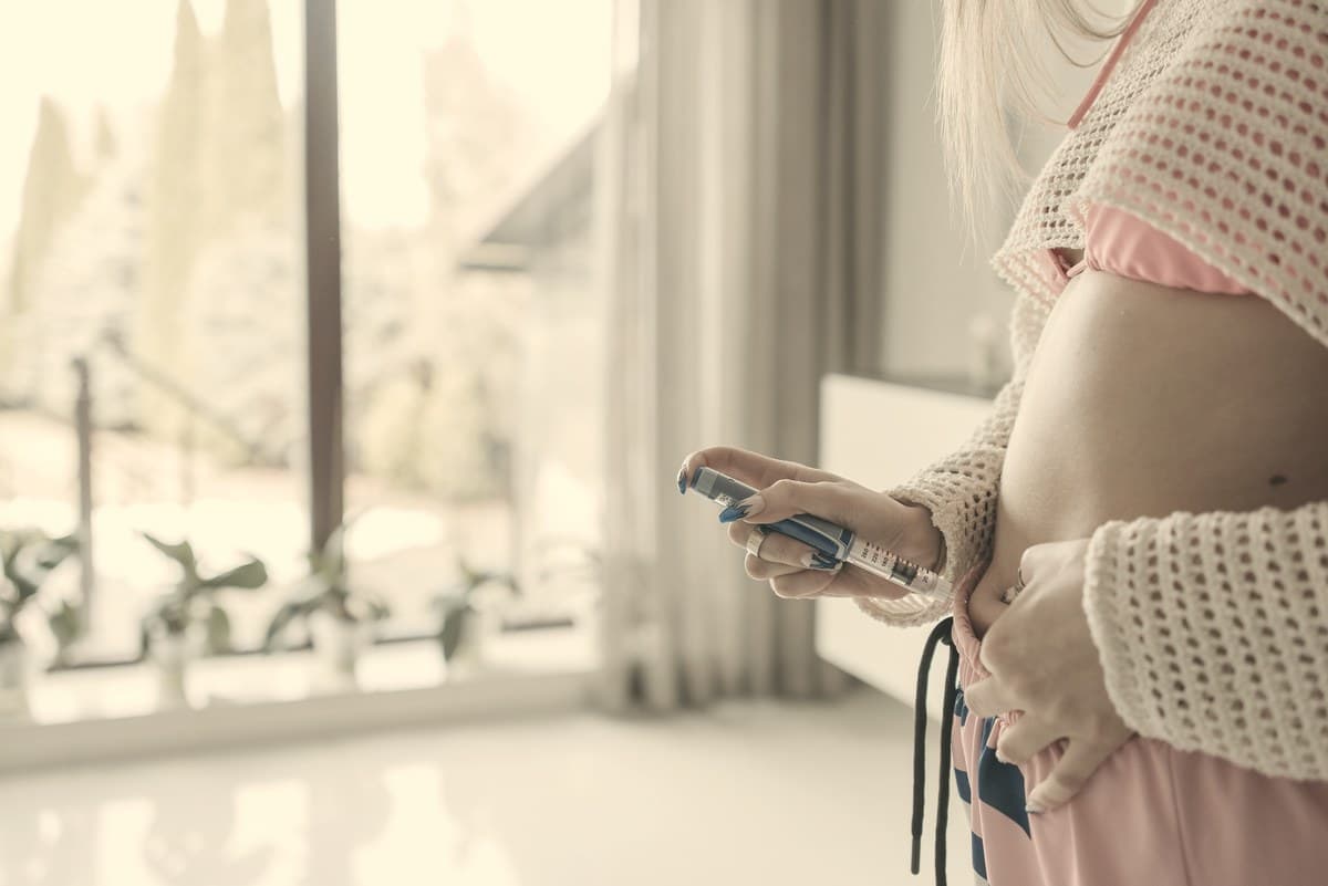 A person holding an injection pen beside their abdomen in soft window light. The abdomen is one of three injection sites the Wegovy label lists.