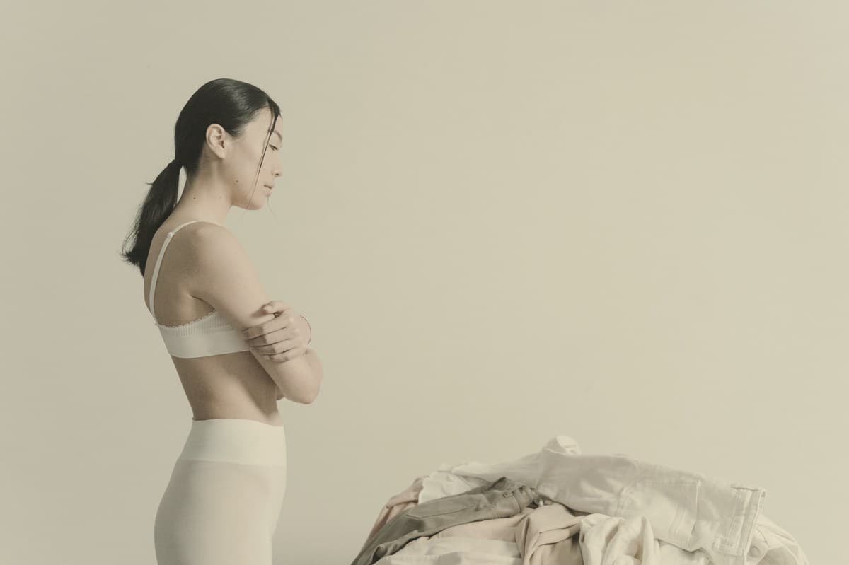 A woman in a white sports top standing in profile with her arms crossed, in soft even light.