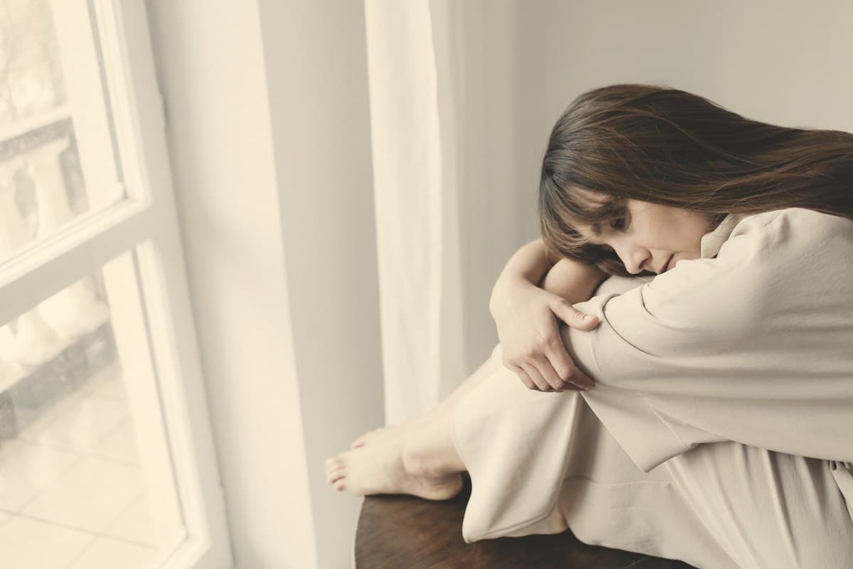A woman sitting by a window, arms folded over her knees, looking unwell in soft natural light.