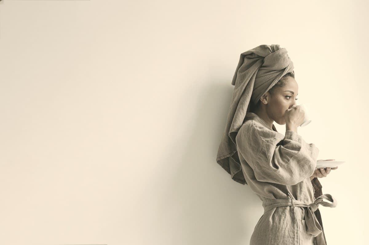 A woman in a robe sipping from a cup against a plain neutral wall in soft morning light.