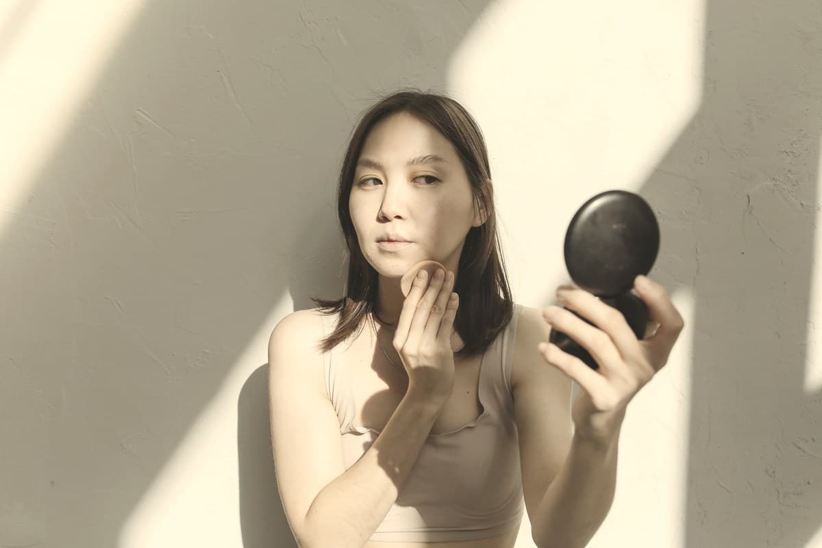 A woman checking her face in a small hand mirror against a sunlit neutral wall.