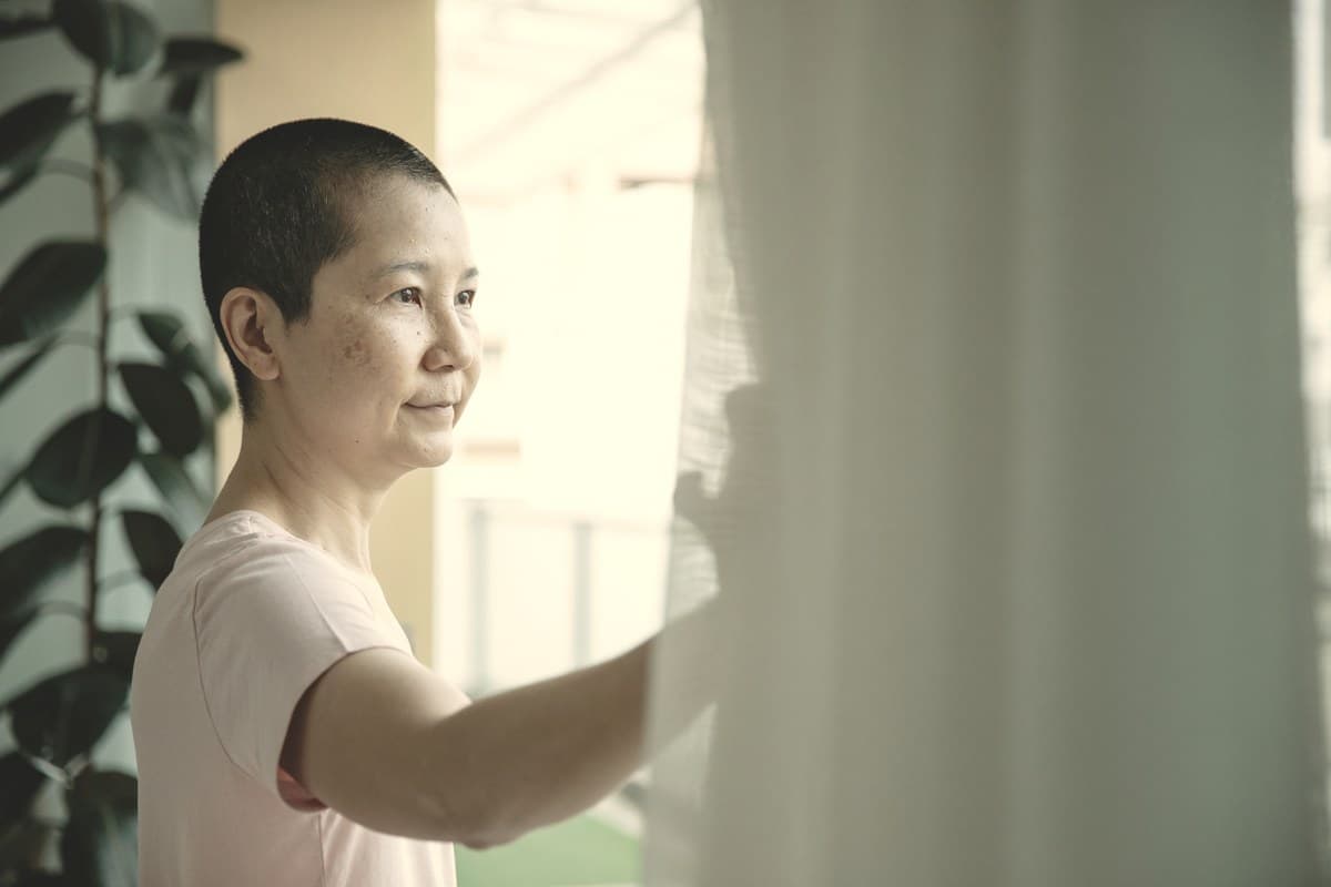 A woman looking out a window in soft daylight, drawing back a sheer curtain.