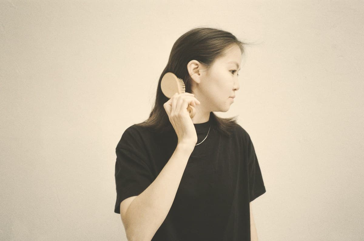 A woman brushing her hair in soft natural light against a plain wall, illustrating hair loss reported by women on Zepbound.